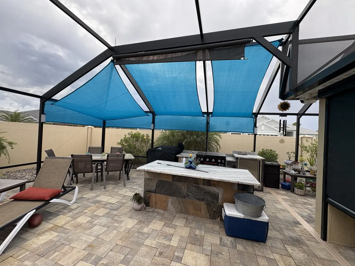 Blue shade sails over an outdoor kitchen island and dining area inside a pool cage